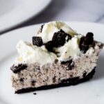 A side view of a slice of homemade oreo pie on a serving plate topped with whipped cream and oreo pieces.