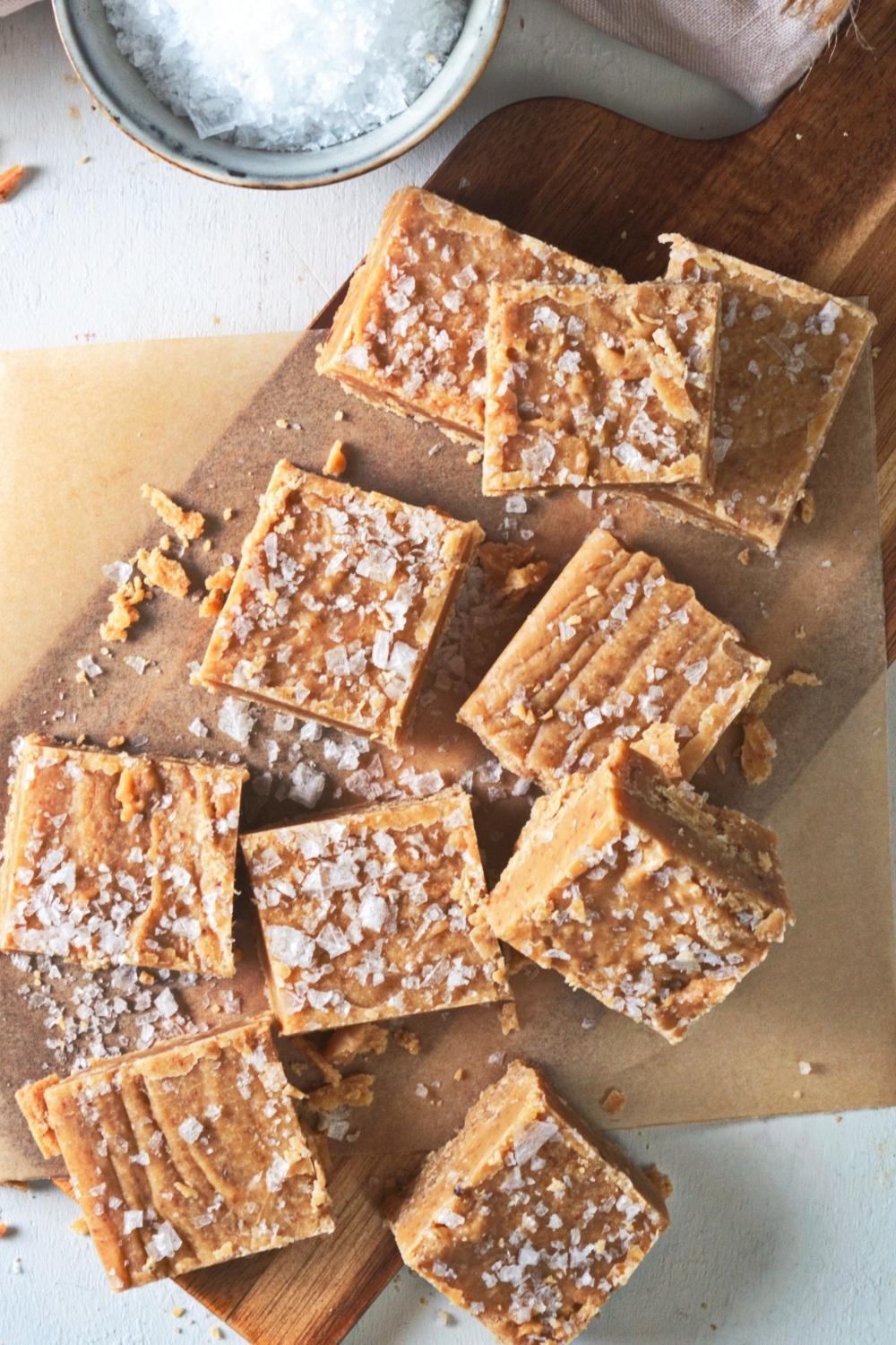 An overhead view of many caramel fudge squares on parchment paper on top of a cutting board with salted caramel on top.