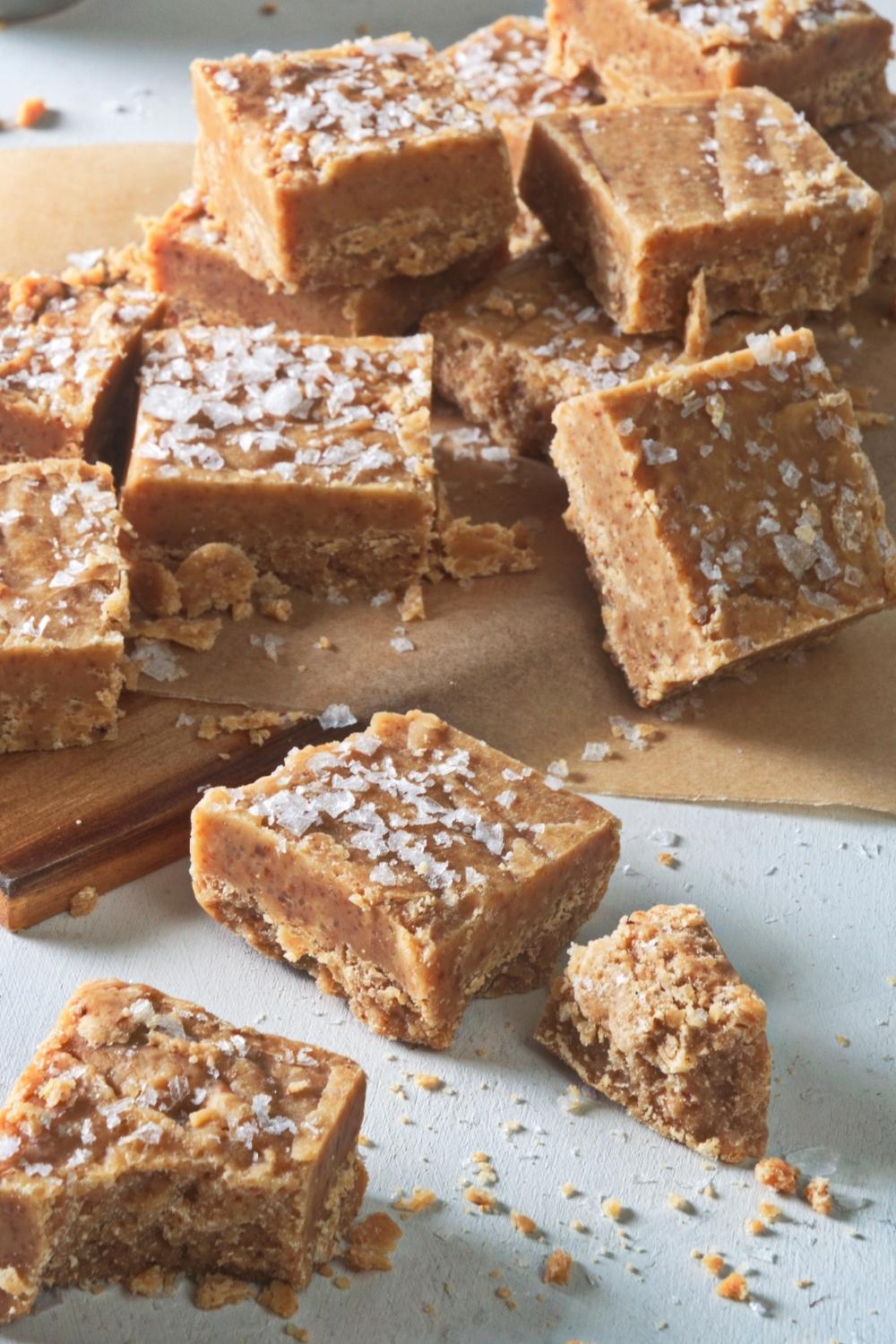 Many caramel fudge squares lay on parchment paper over a cutting board. They are all garnished with salted caramel on top.