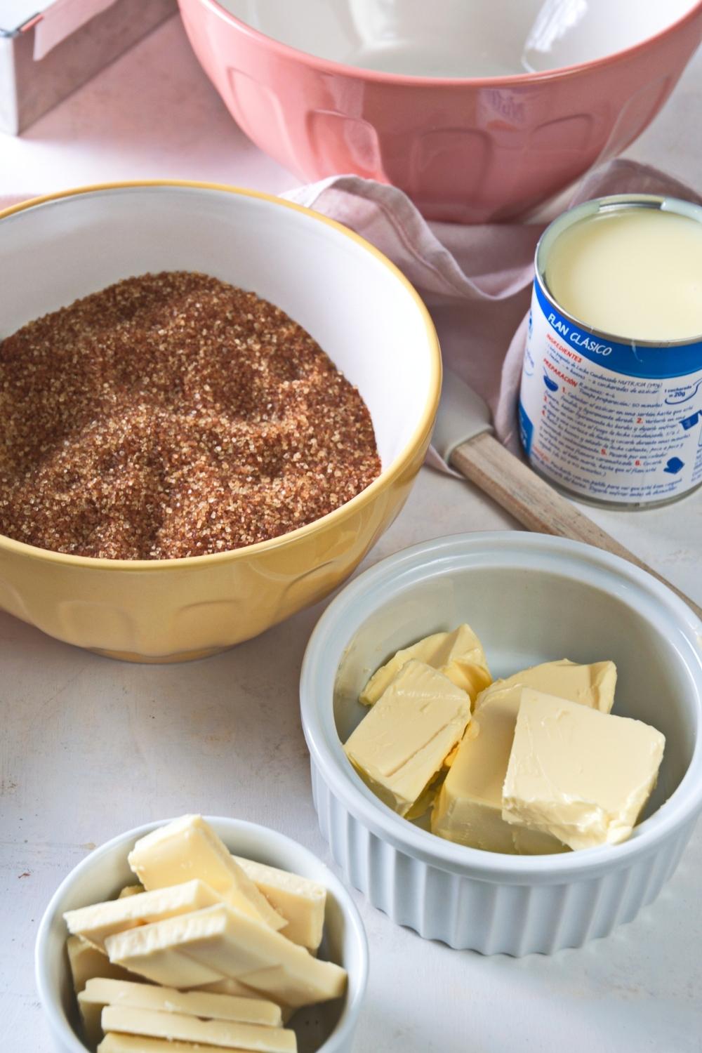A large bowl containing brown sugar, a medium size bowl containing butter, a can of condensed milk, and a small bowl of white chocolate bar chunks.