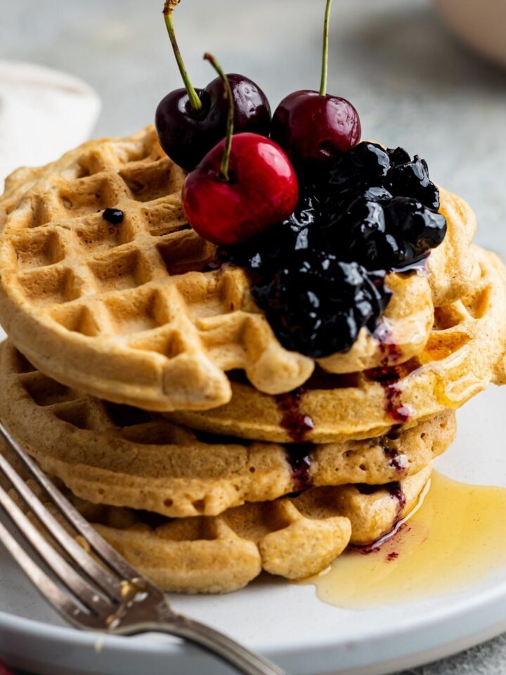 A stack of four homemade waffles without milk, drizzled with honey and topped with fresh berries and cherries.