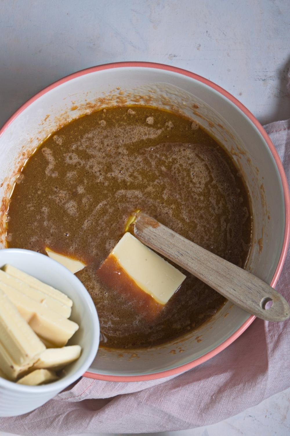 An overhead view of melted caramel fudge ingredients with a spatula and a small bowl containing white chocolate bar chunks being poured into it.