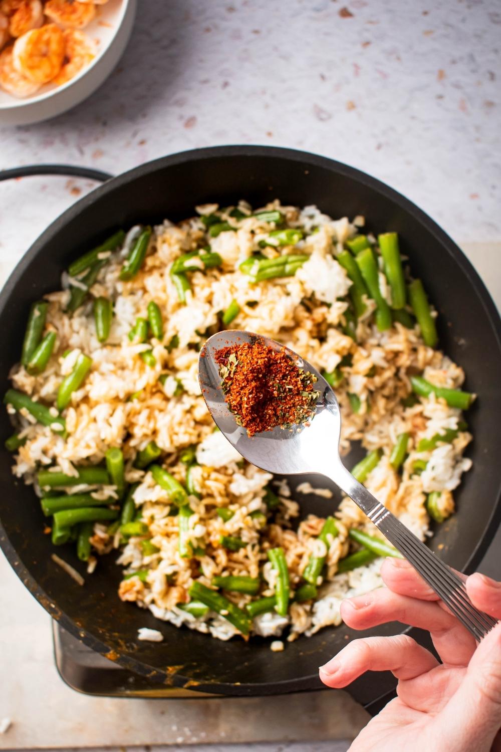 A hand holding a spoon over a skillet filled with rice and cut green beans.