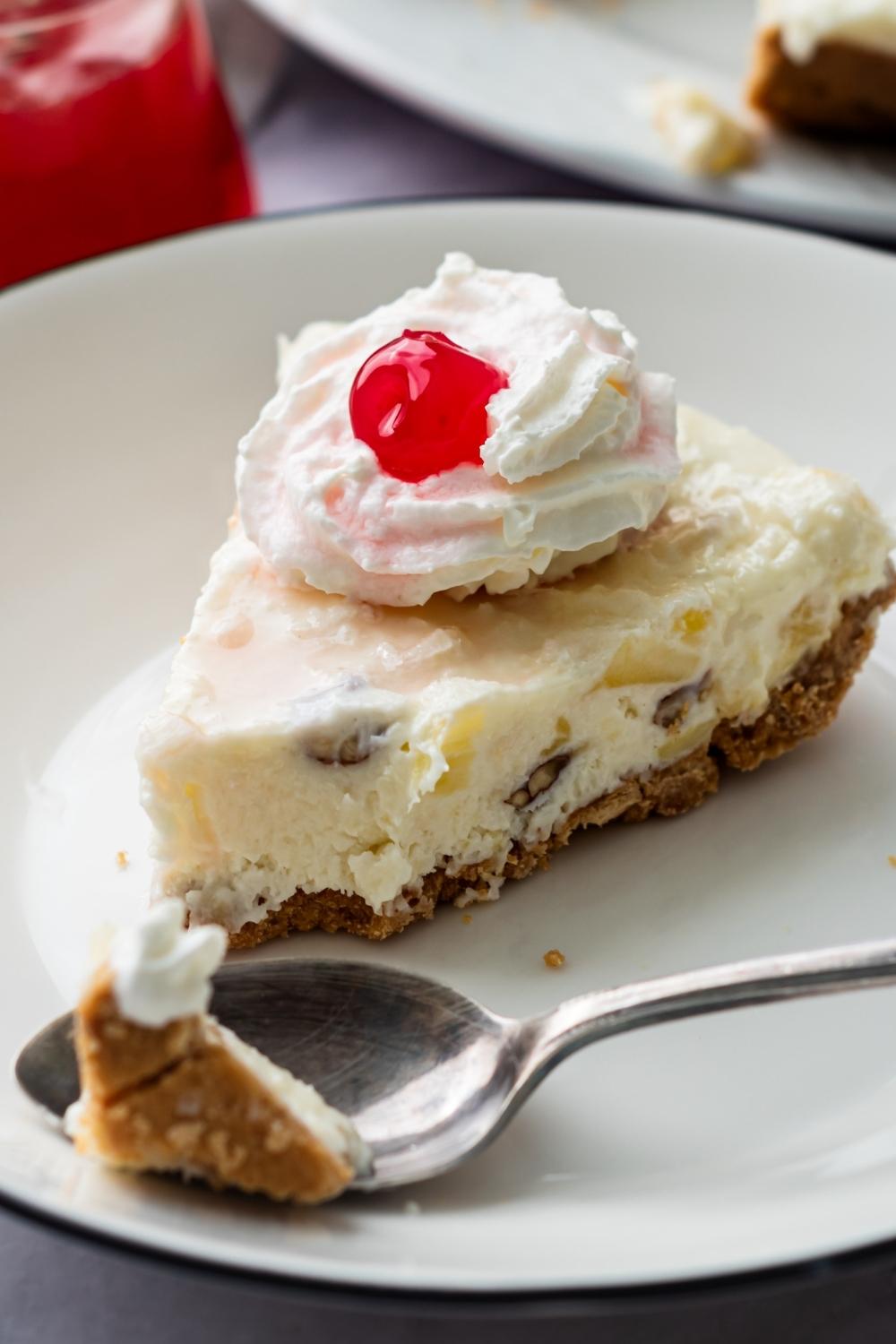 A close up of a serving dish with 1 slice of million dollar pie topped with whipped cream and a cherry. A spoon has a piece of million dollar pie in it.