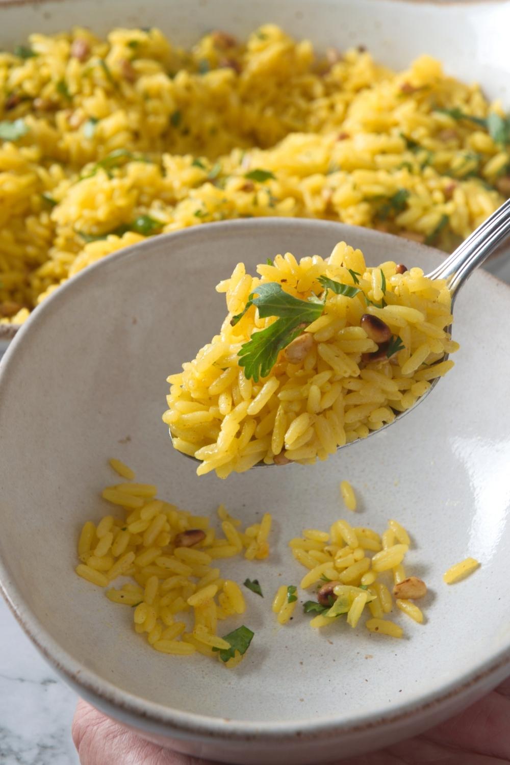 A spoon with yellow rice on it hovering over an empty bowl. Behind it is more yellow rice.