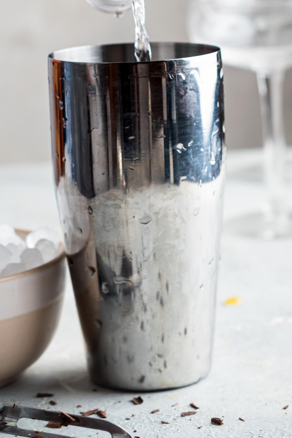 A profile view of a stainless steel cocktail shaker with condensation on the sides and a shot glass full of vodka being poured into it.