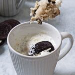 A close-up of a mug containing homemade Oreo mug cake topped with whipped cream and a half an Oreo. A spoon holds a heaping scoop of the homemade Oreo mug cake above the mug.