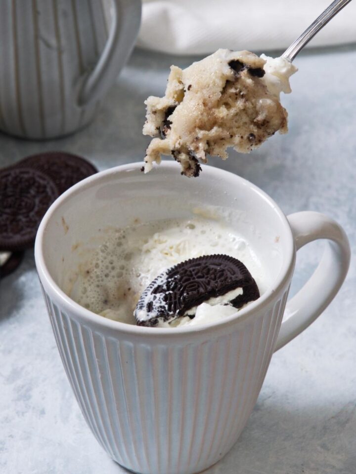 A close-up of a mug containing homemade Oreo mug cake topped with whipped cream and a half an Oreo. A spoon holds a heaping scoop of the homemade Oreo mug cake above the mug.