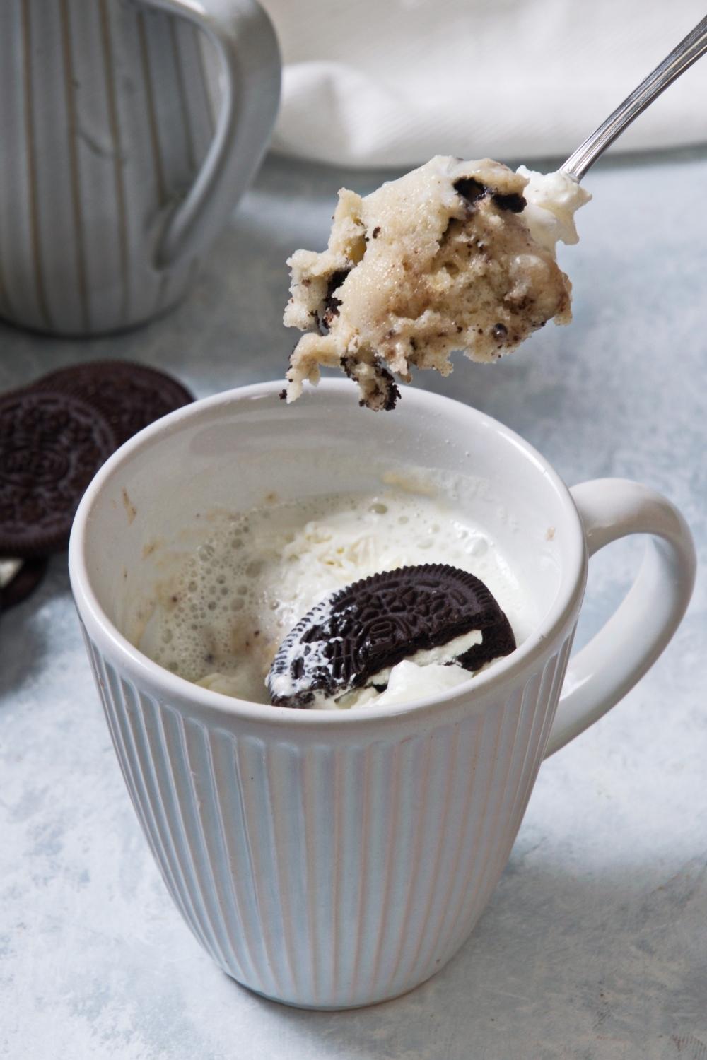 A close-up of a mug containing homemade Oreo mug cake topped with whipped cream and a half an Oreo. A spoon holds a heaping scoop of the homemade Oreo mug cake above the mug.