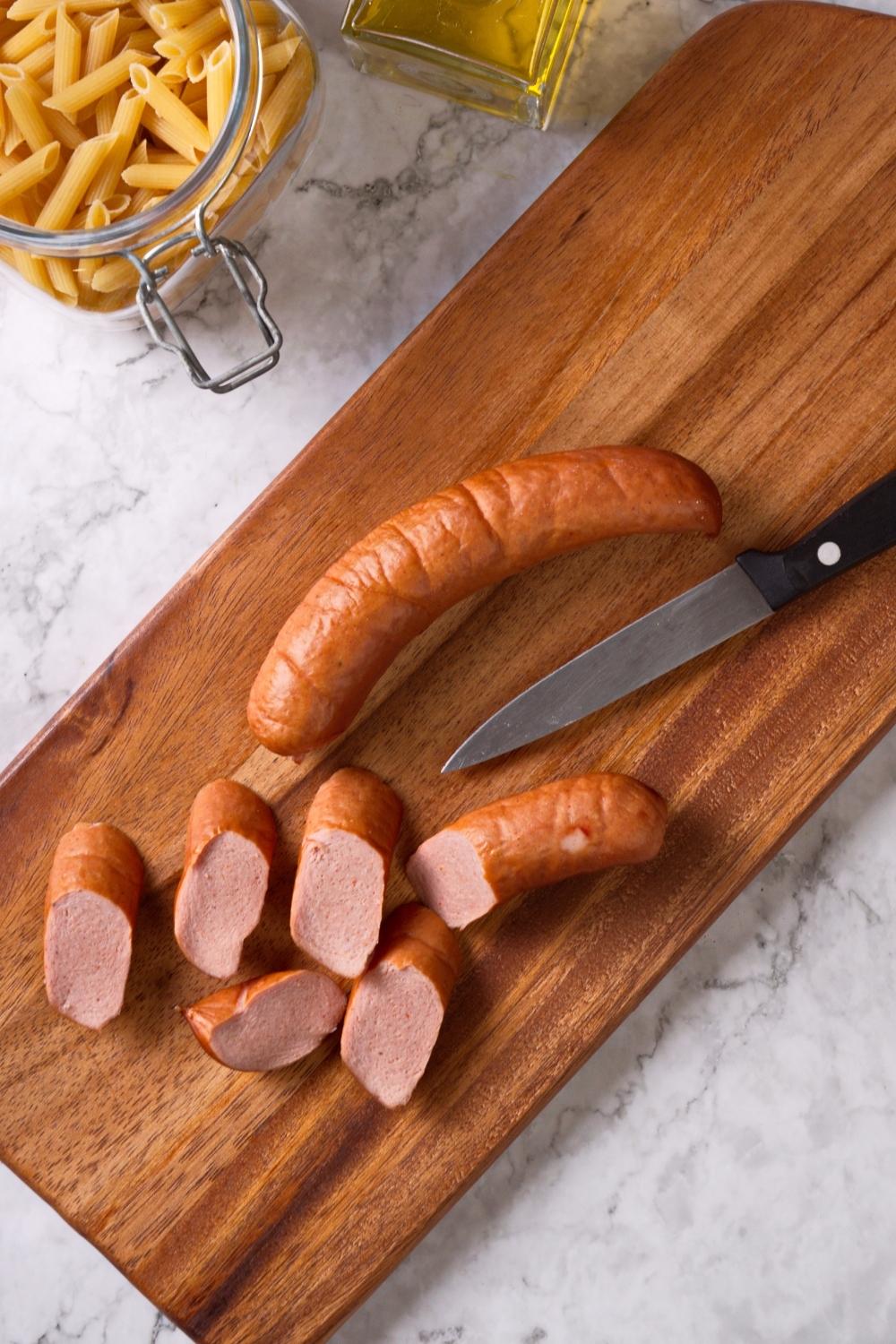 An overhead view of sausage on a cutting board. Half of it is cut into chunks the other half is not. The knife sets next to the sausage.