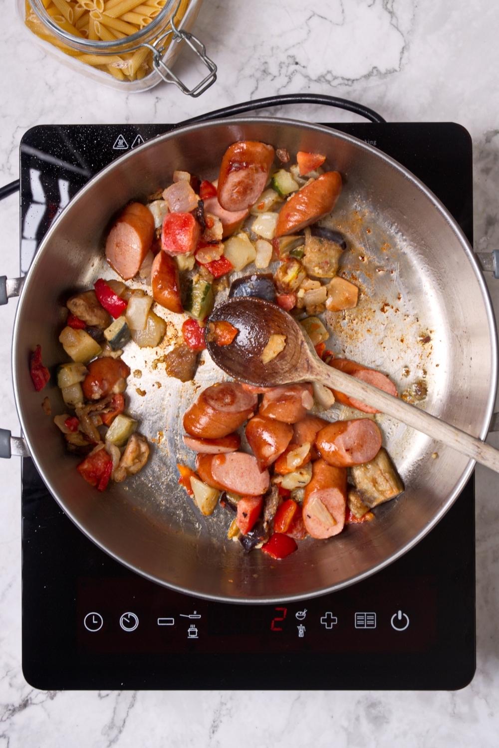 An overhead view of a cooking pan cooking mixed veggies and chunks of sausage.