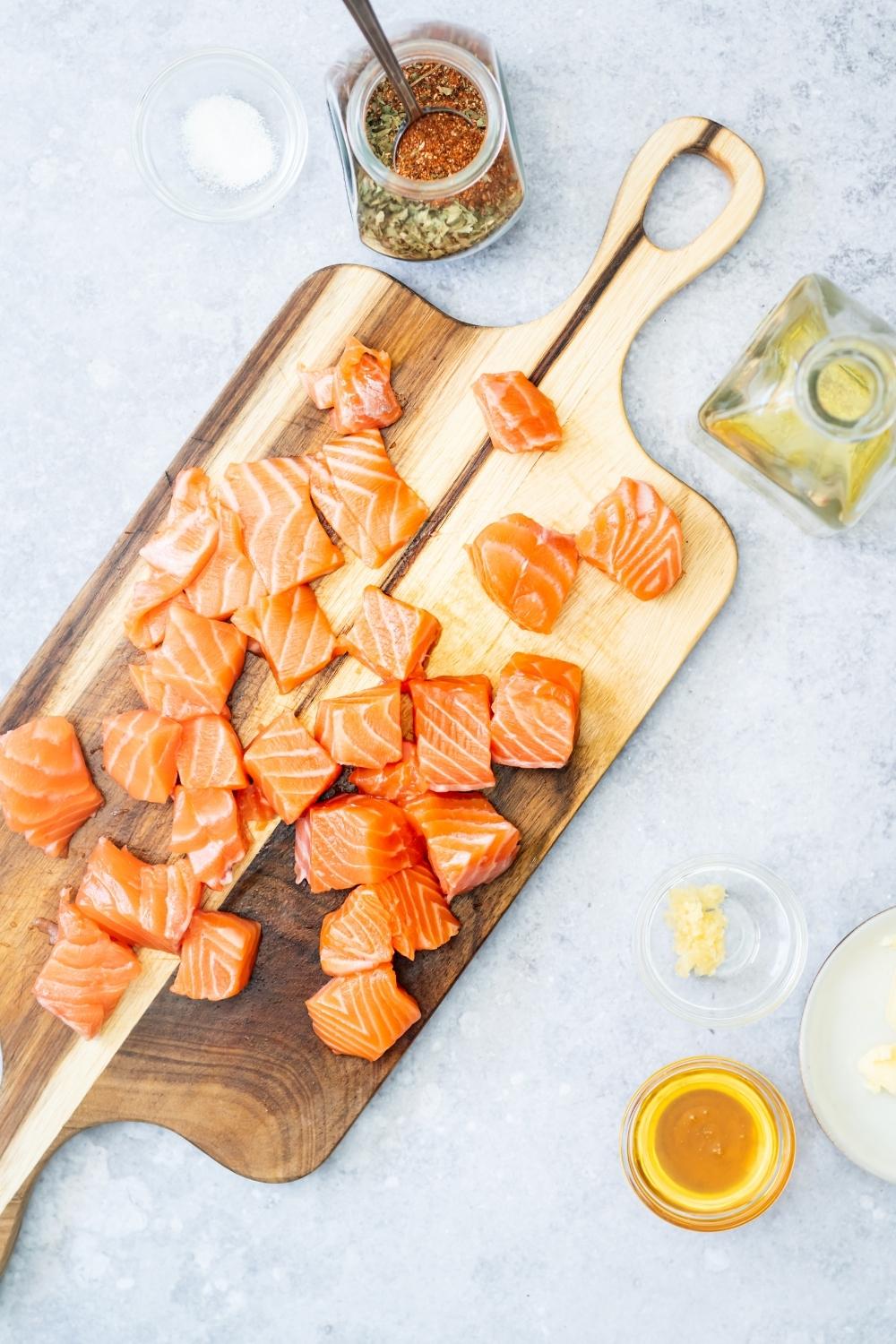 Salmon fillet pieces on a wooden cutting board on a grey counter. Behind the cutting board is a glass jar filled with blackened seasonings.