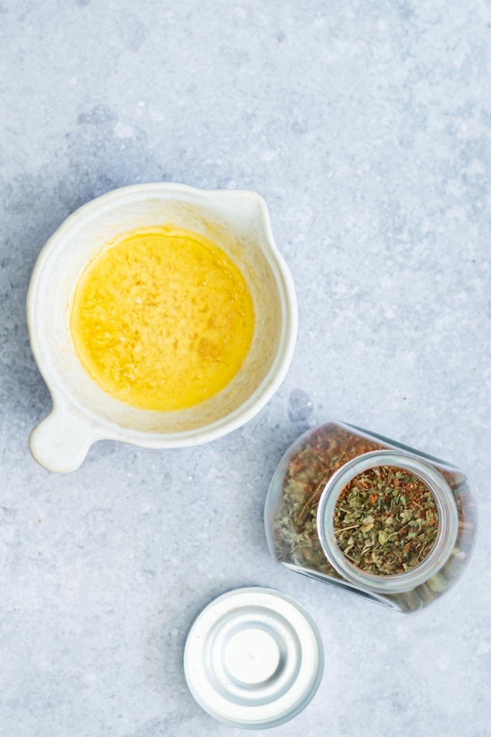 A bowl of melted butter and a glass jar of blackened seasoning on a grey counter.