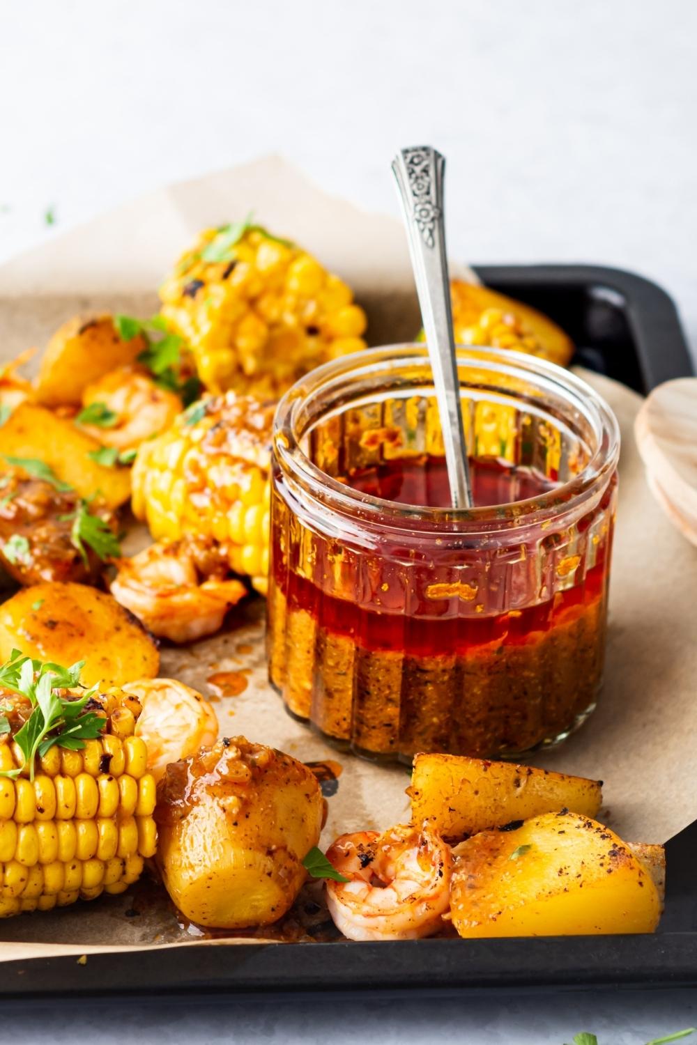 A jar containing cooked seafood boil sauce with a spoon in it. It is surrounded by cooked veggies and shrimp.