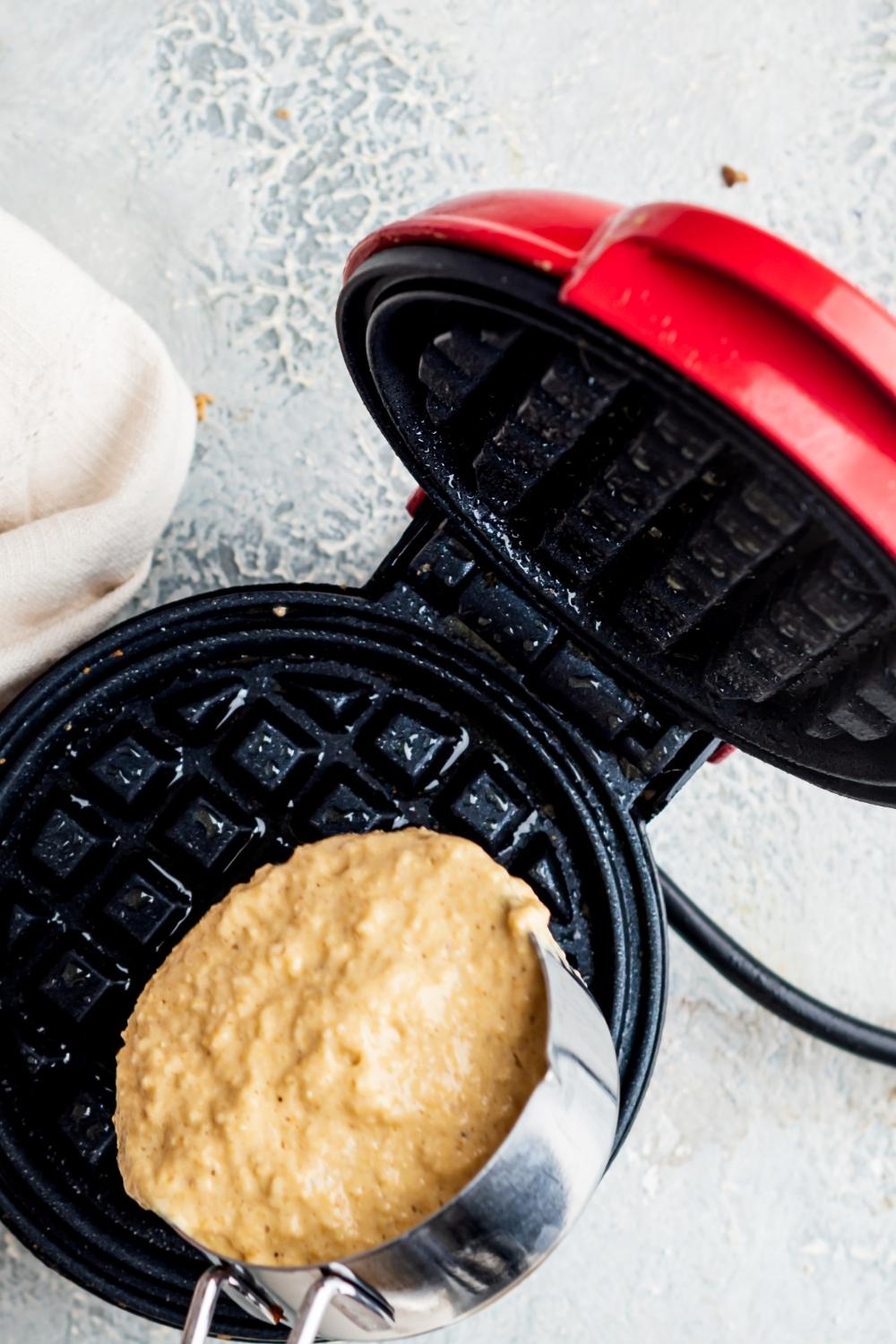 An overhead view of a personal size waffle maker and a measuring cup pouring waffle batter into the hot waffle iron.