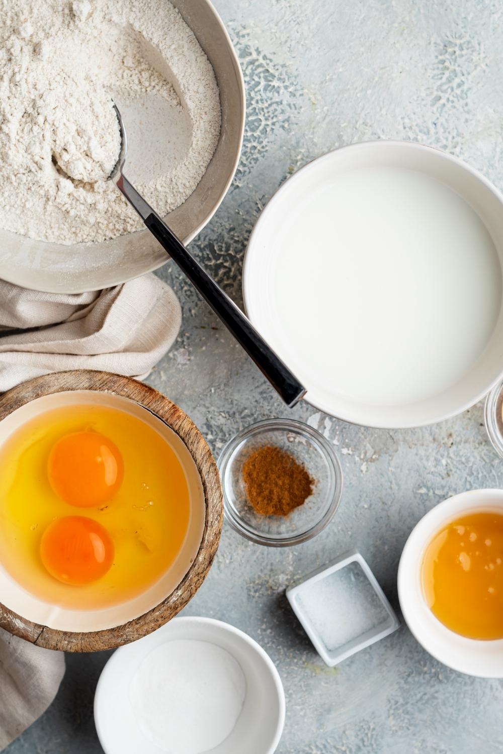 An overhead view of mixing bowls containing oat flour, almond milk, two eggs cracked in a bowl, cinnamon, salt, honey, and baking powder.