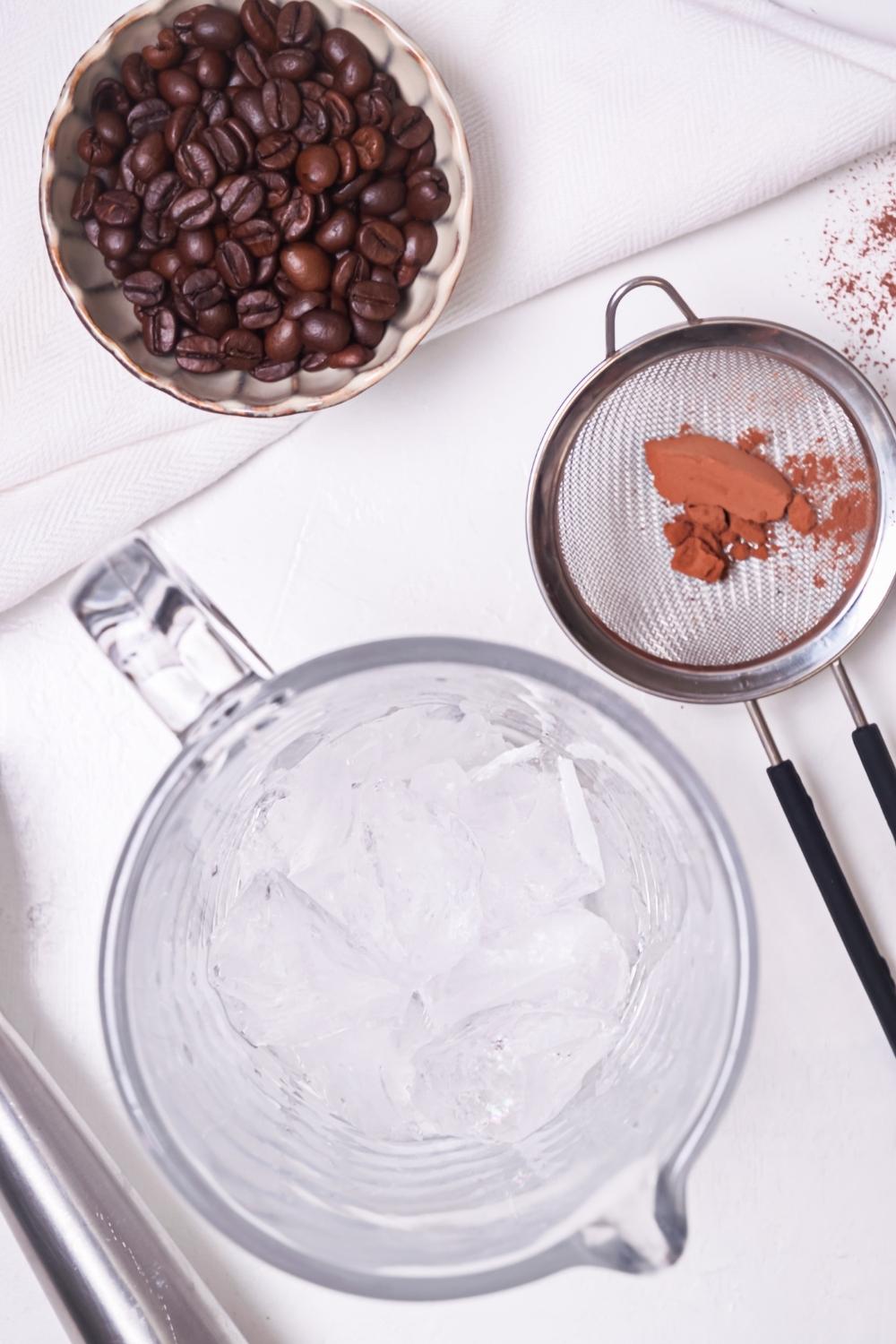 An overhead view of a tall glass of ice, a sister with cinnamon, and coffee beans in a small bowl.