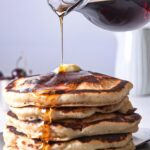 A stack of homemade McDonald's pancakes on a plate topped with butter; maple syrup is being poured on them.