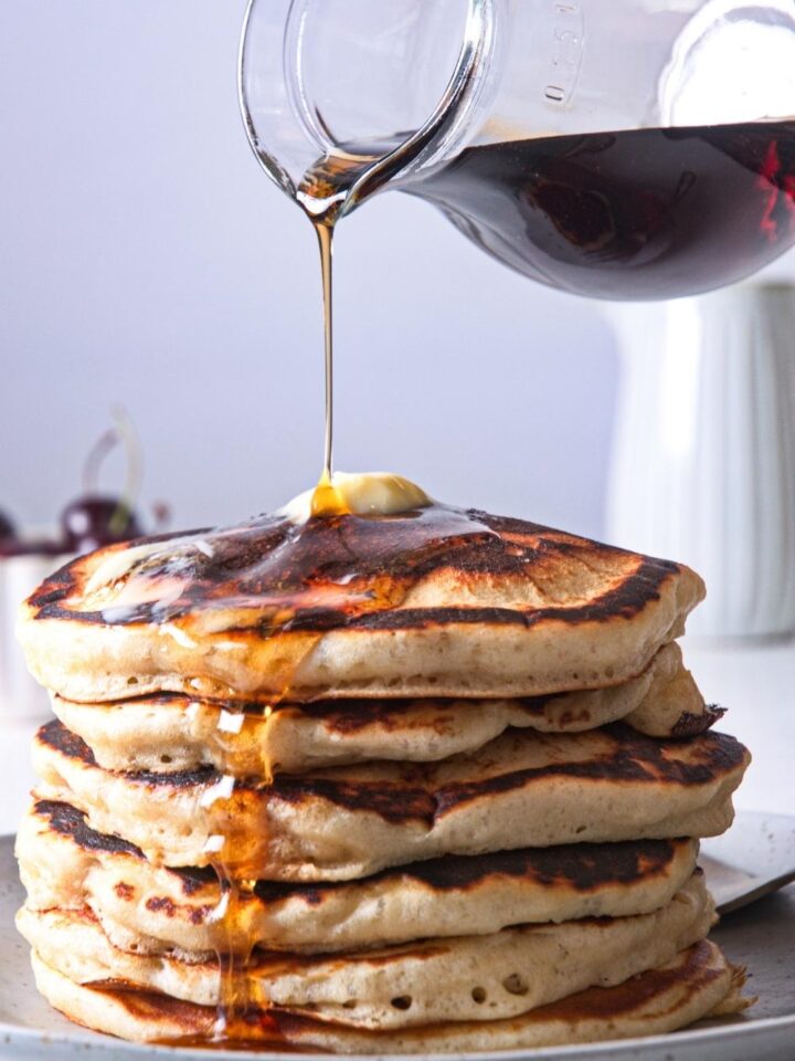 A stack of homemade McDonald's pancakes on a plate topped with butter; maple syrup is being poured on them.