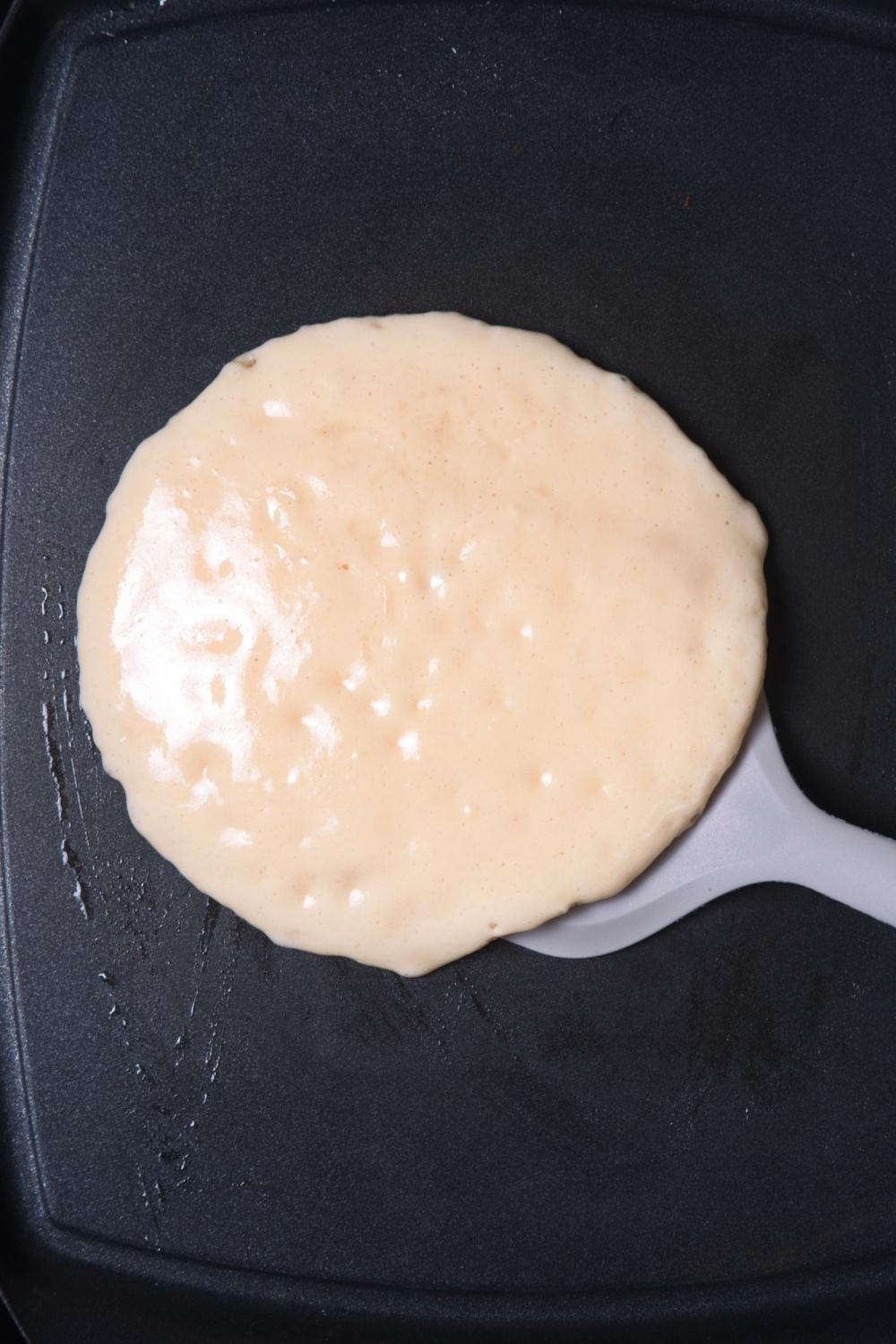 A half cooked pancake being flipped by a spatula on a hot griddle.