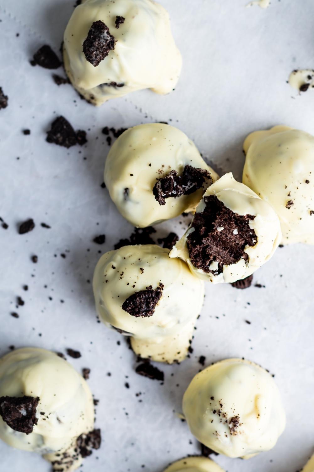 An overhead view of oreo balls on parchment paper. Each oreo ball is dipped in white chocolate and topped with crumbled oreos. One oreo ball has a bite taken out of it.