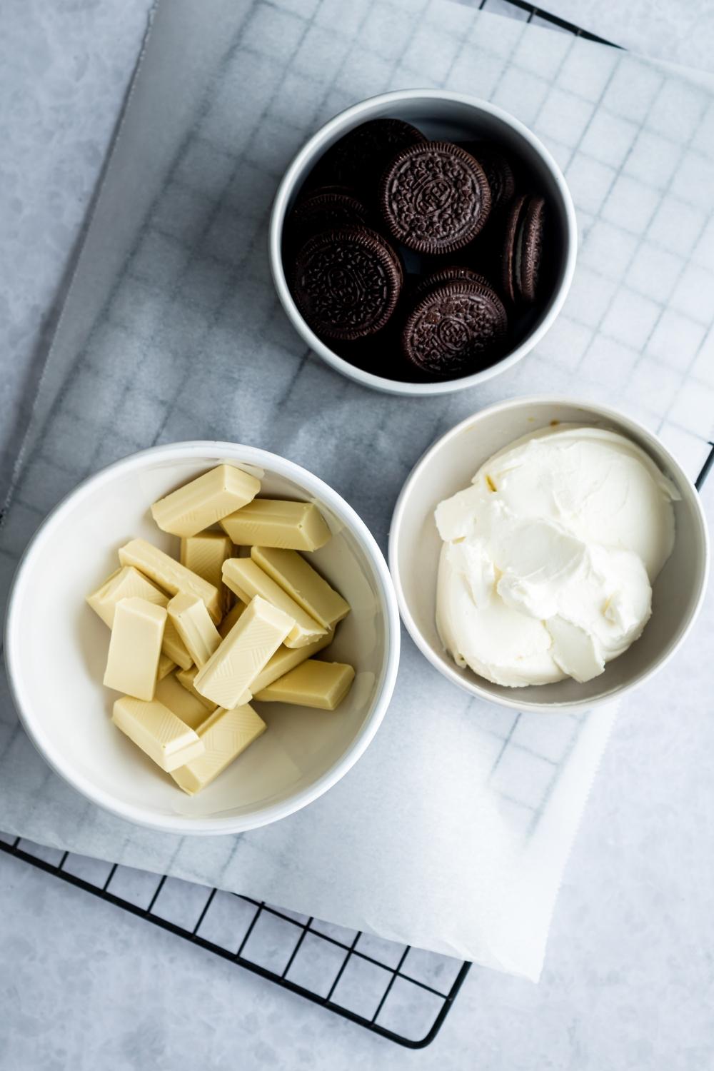 An overhead view of 3 small bowls containing white chocolate melting bars, cream cheese, and oreos.