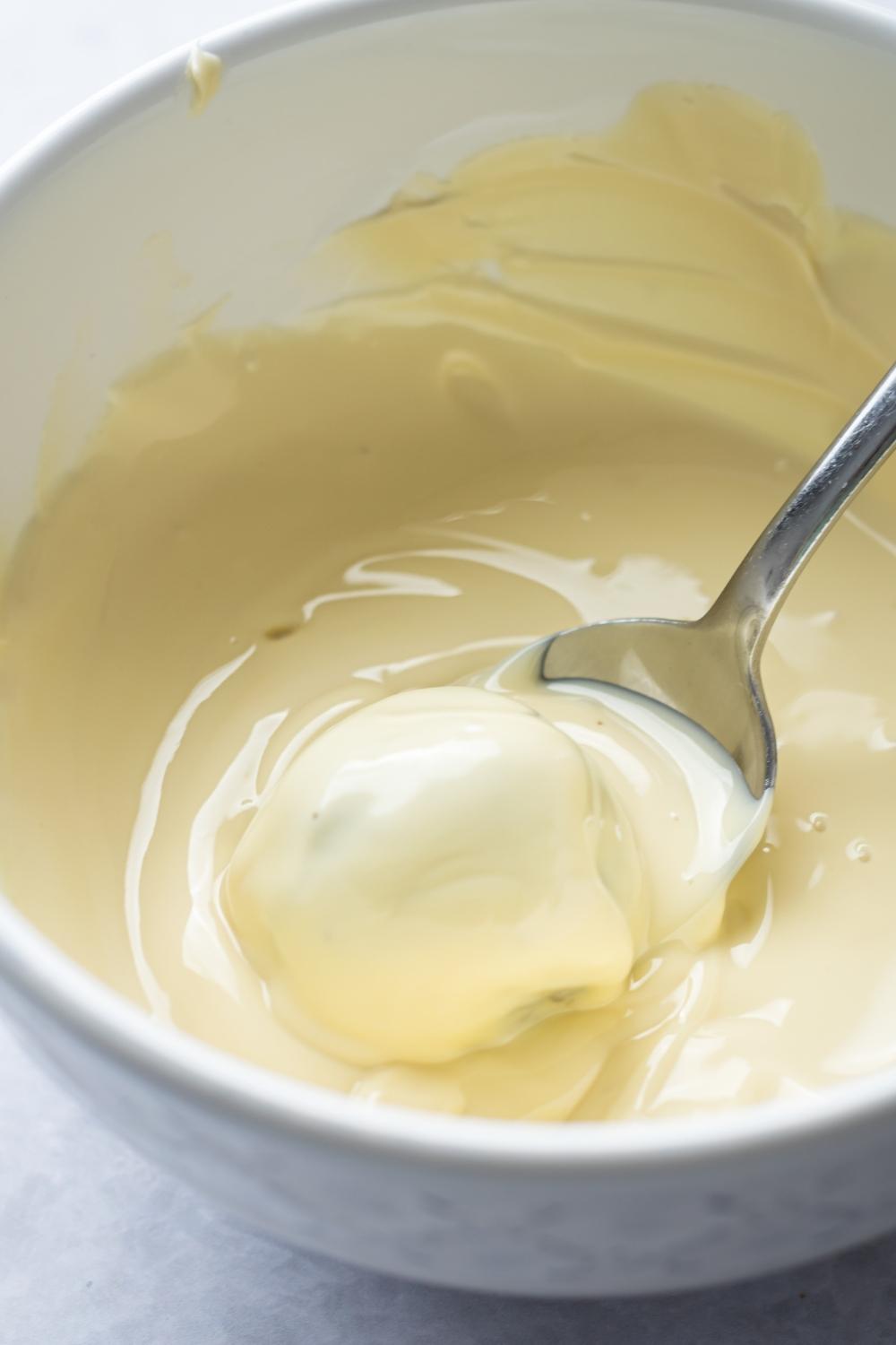 A close up of a bowl with melted white chocolate. A spoon is holding an oreo and dipping it in completely.