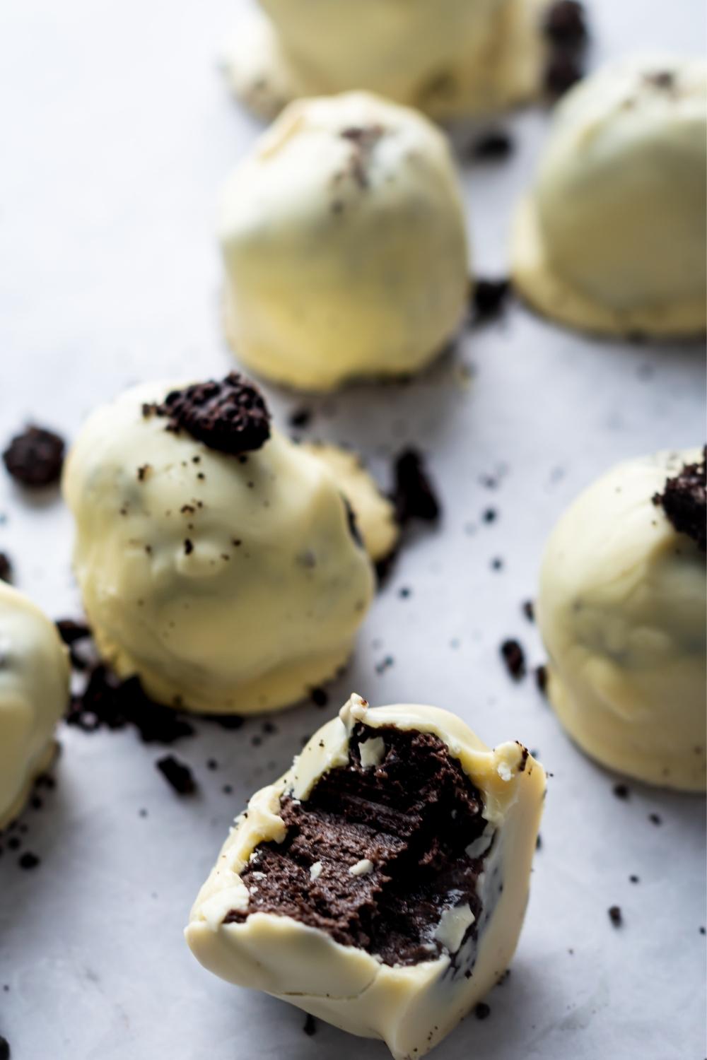 A close up of oreo balls on parchment paper. Each oreo ball is dipped in white chocolate and topped with crumbled oreos. One oreo ball has a bite taken out of it.