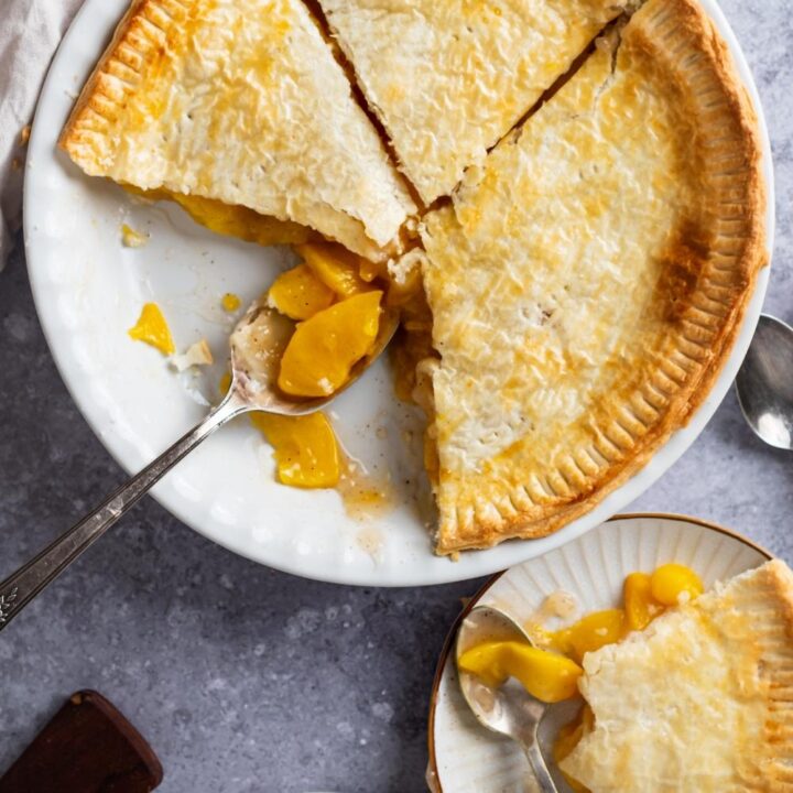 View of a pie dish with baked peach pie. Two slices have been taken out. A spoon sets with peaches in it where the slices ones were.