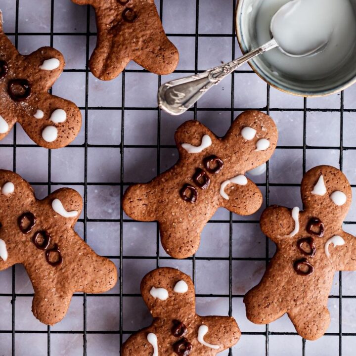 An overhead view of decorated gingerbread men on a wire rack. A small bowl of homemade icing with a spoon in it set aside.