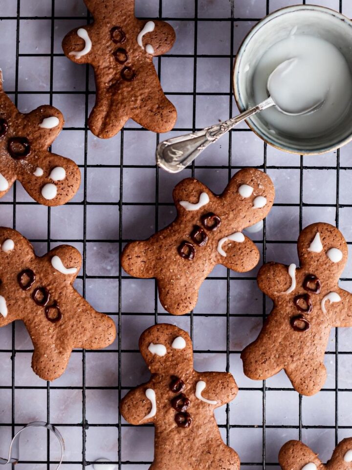 An overhead view of decorated gingerbread men on a wire rack. A small bowl of homemade icing with a spoon in it set aside.