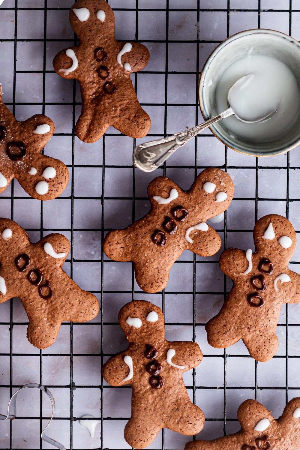 An overhead view of decorated gingerbread men on a wire rack. A small bowl of homemade icing with a spoon in it set aside.