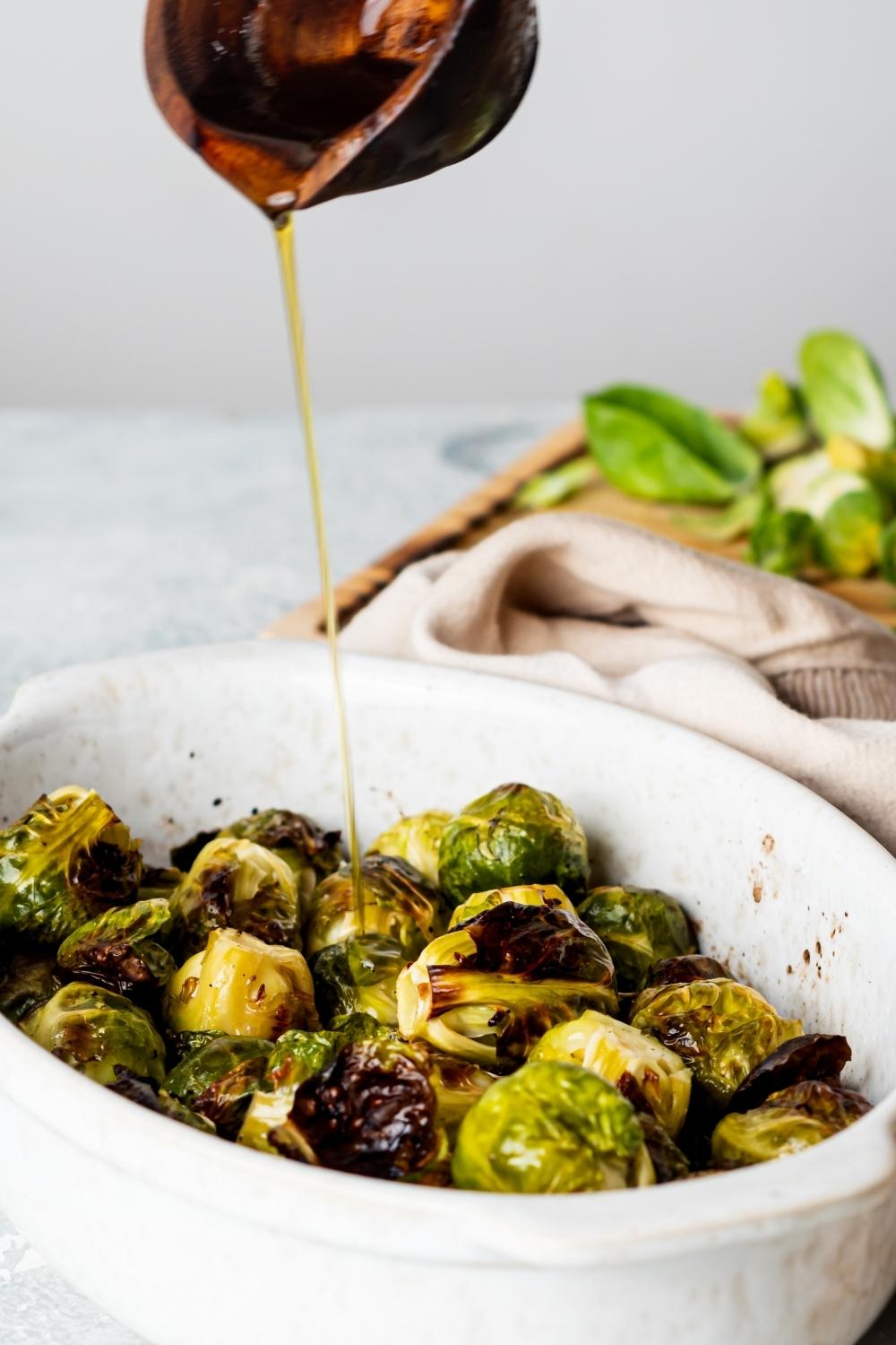 A casserole dish with cooked brussels sprouts. A small spouted cup s pouring honey onto the sprouts.