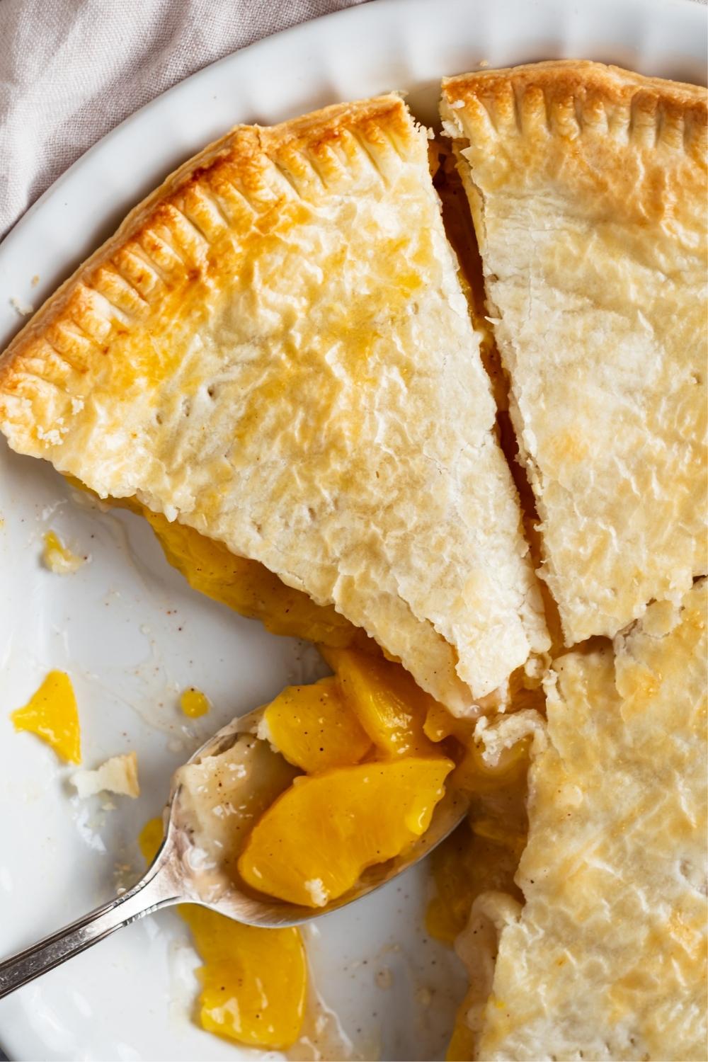A pie dish with baked peach pie. Two slices have been taken out. A spoon sets with peaches in it where the slices ones were.