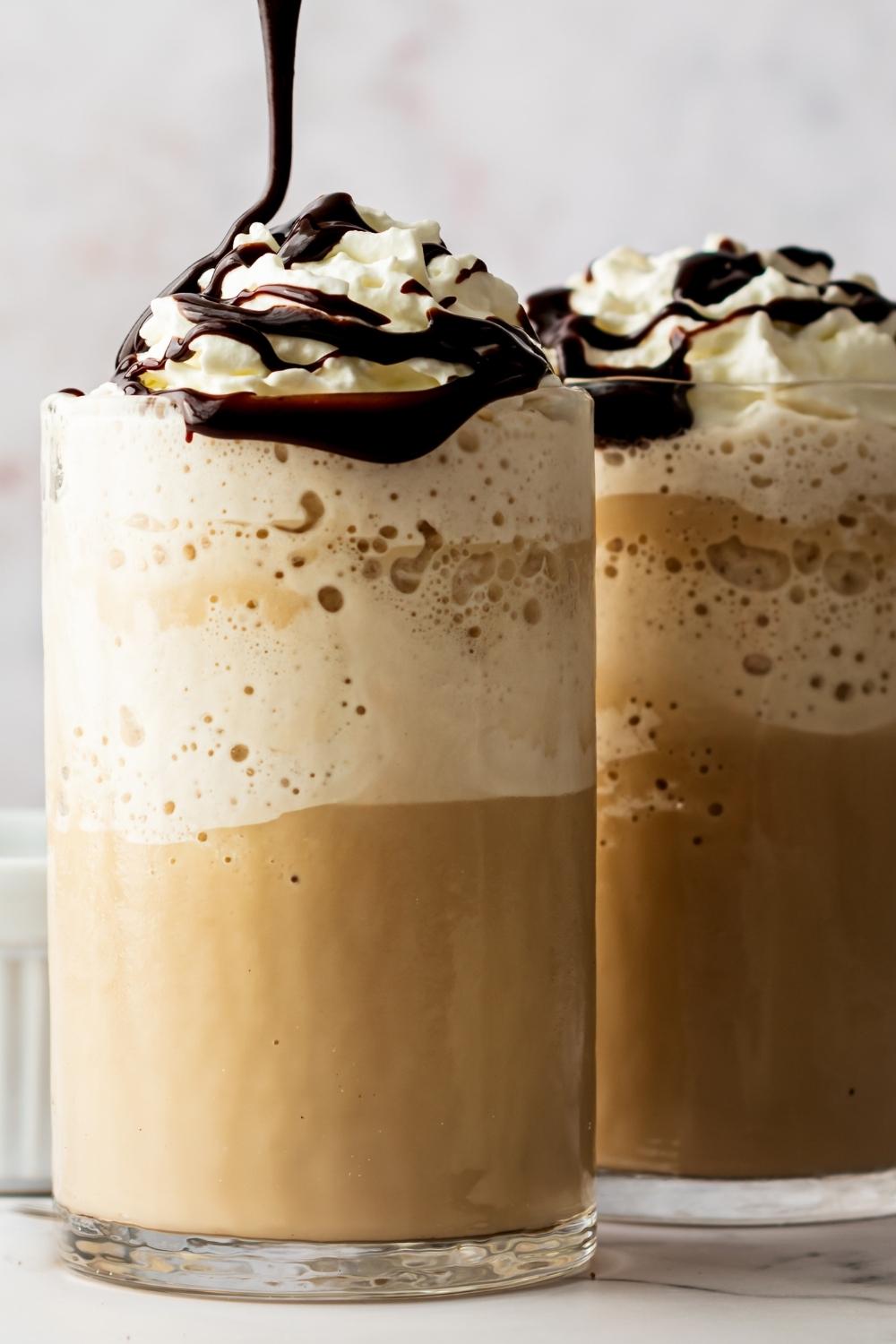 Two glasses of homemade McDonald's mocha frappe topped with whipped cream. A chocolate drizzle is drizzling over top of the whipped cream.