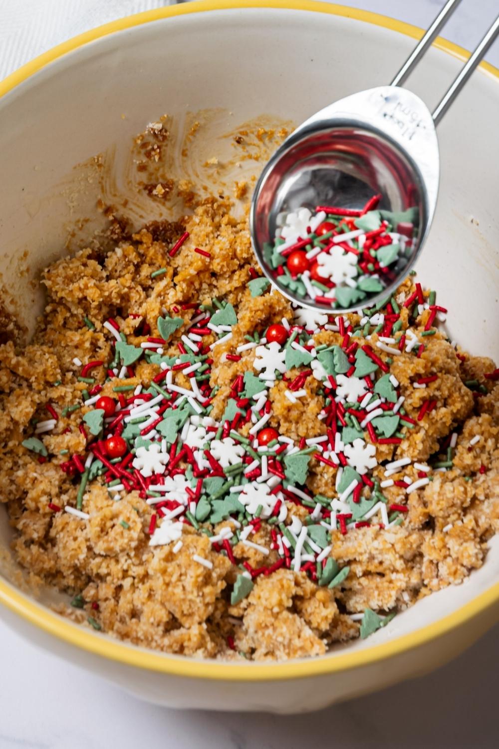 A measuring cup with christmas sprinkles being held over a bowl with the sprinkles on top of a no bake peanut butter cookie dough.