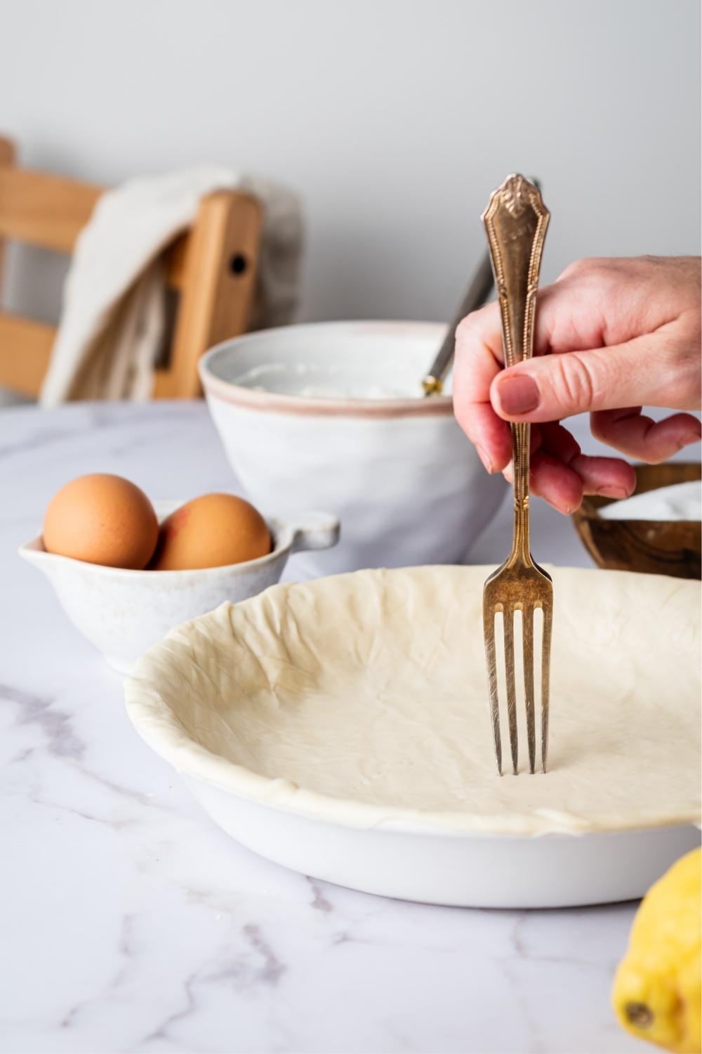 A hand piercing holes in a pie crust in a pie dish. Behind it is two eggs in a bowl and a bowl of ricotta cheese.