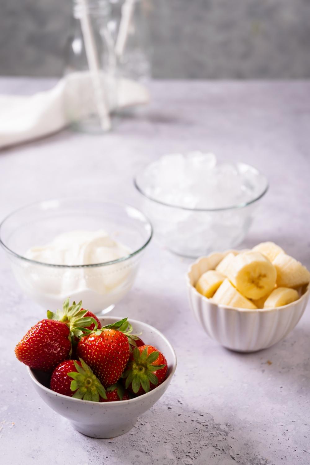 A bowl of strawberries, a bowl of sliced bananas, a bowl of yogurt, and a bowl of ice on a counter.