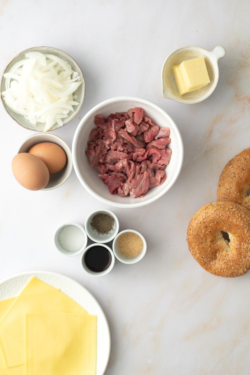 A bowl of shaved steak, a bowl with two eggs in it, a bowl of sliced onions, a bowl of butter, part of a plate with cheese slices, and part of two bagels all on a white counter.