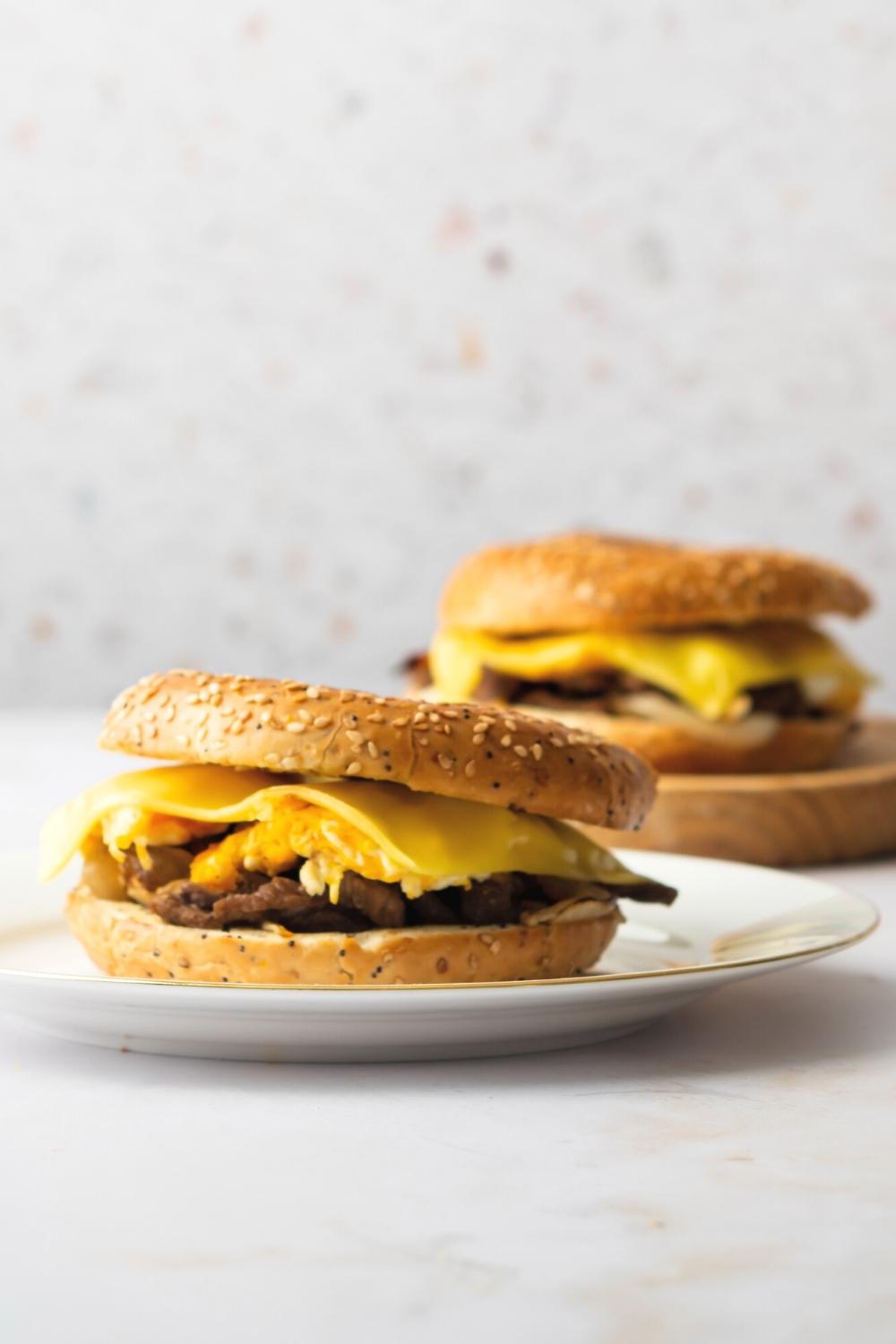 A steak, egg, and cheese bagel sandwich on a white plate. Behind it is the same sandwich on a wood board.