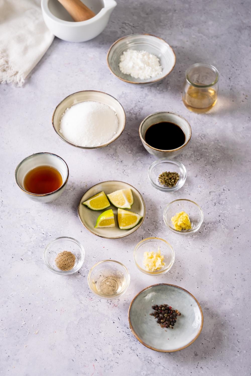 A small plate with szechuan seeds on it, a small bowl of pepper, a small plate with limes, a small bowl of soy sauce, a bowl of sugar, and a bowl of vinegar all on a grey counter.