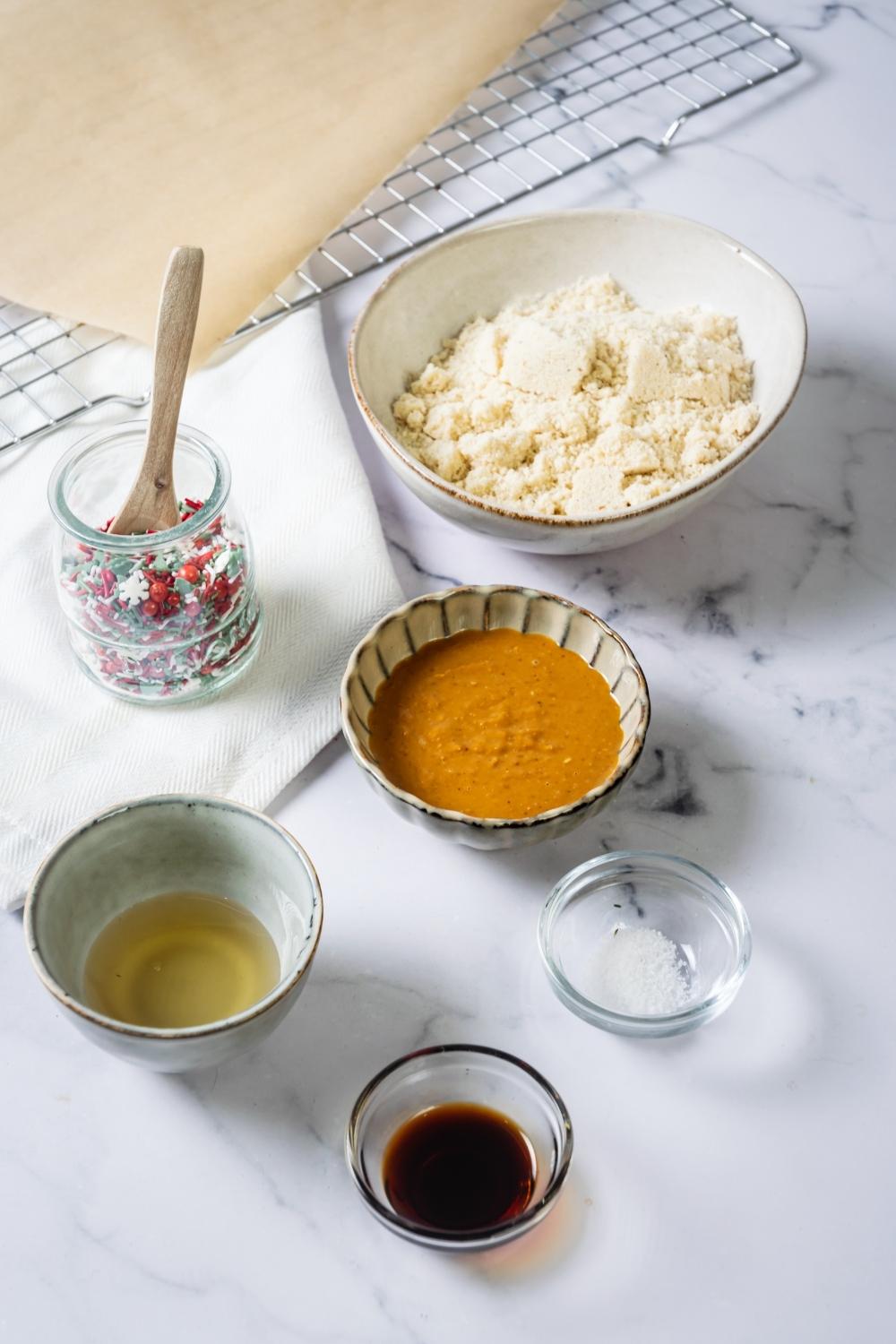 A bowl of flour, a bowl of christmas sprinkles, a bowl of agave, a bowl of salt, and a bowl of vanilla extract all on a grey counter.