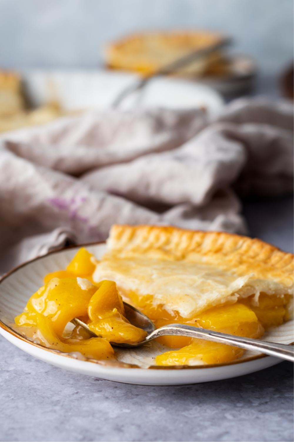 A serving plate with one slice of peach pie that is half eaten. A spoon sets with peach pie filling on it.
