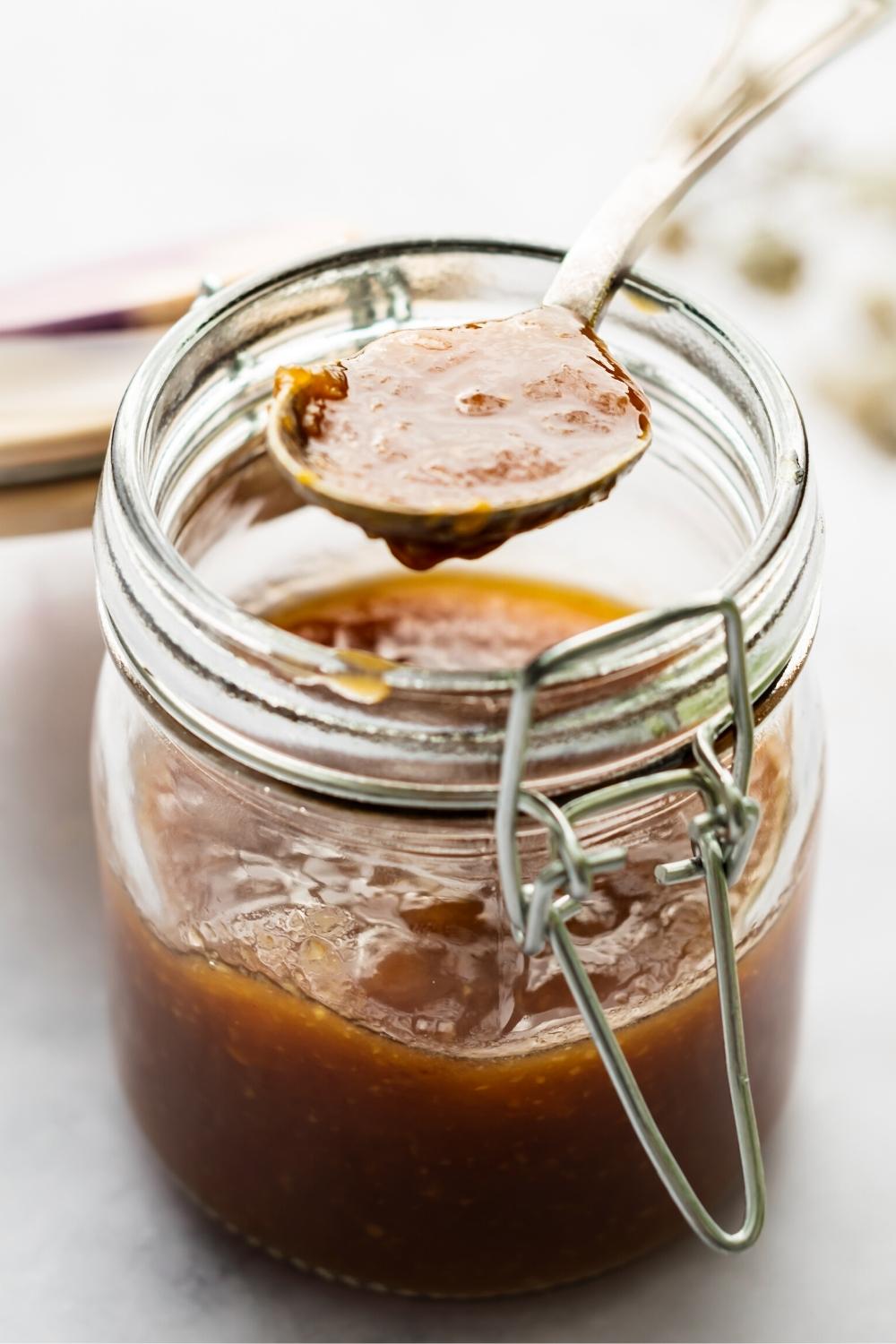A spoon that has sweet and sour sauce on it over the top of a glass jar with the sauce in it.