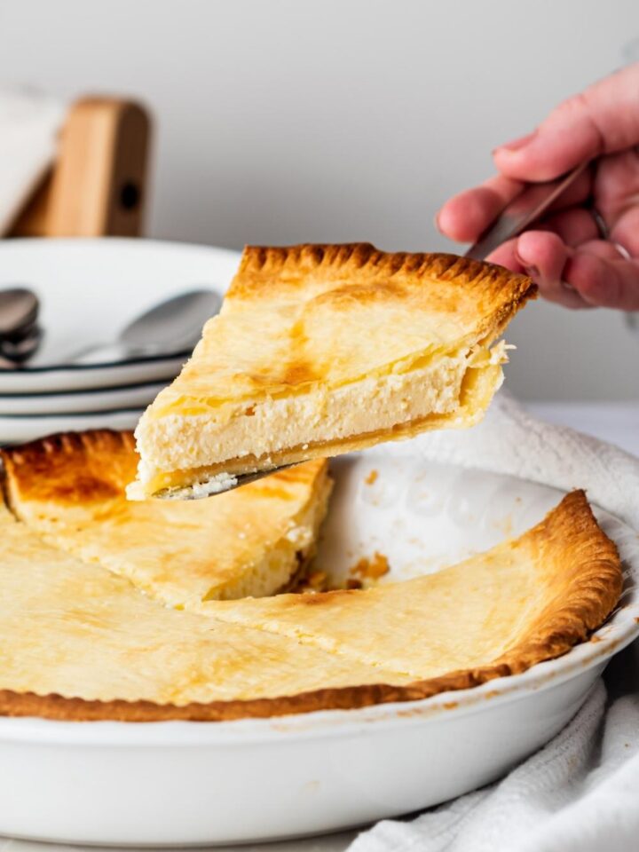 A hand holding a slice of ricotta pie over the whole ricotta pie.