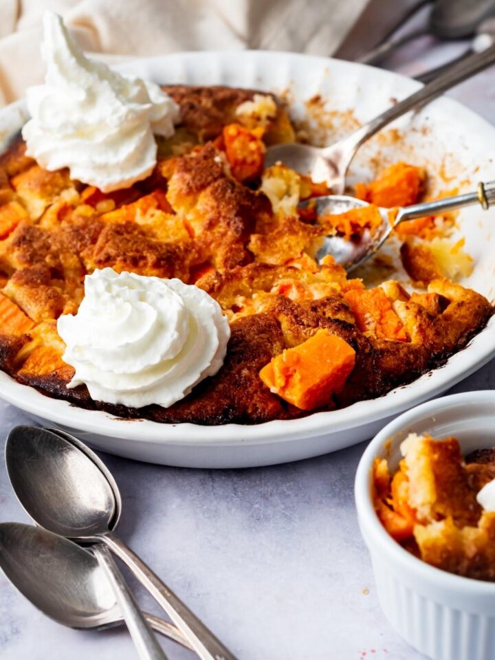A white pie dish with sweet potato cobbler in it. There are two dollops of whipped cream on part of it and there is some cobbler missing on the right side.