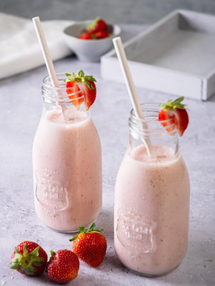 Two strawberry banana smoothies in glasses with a straw in them and sliced strawberry on the rim.