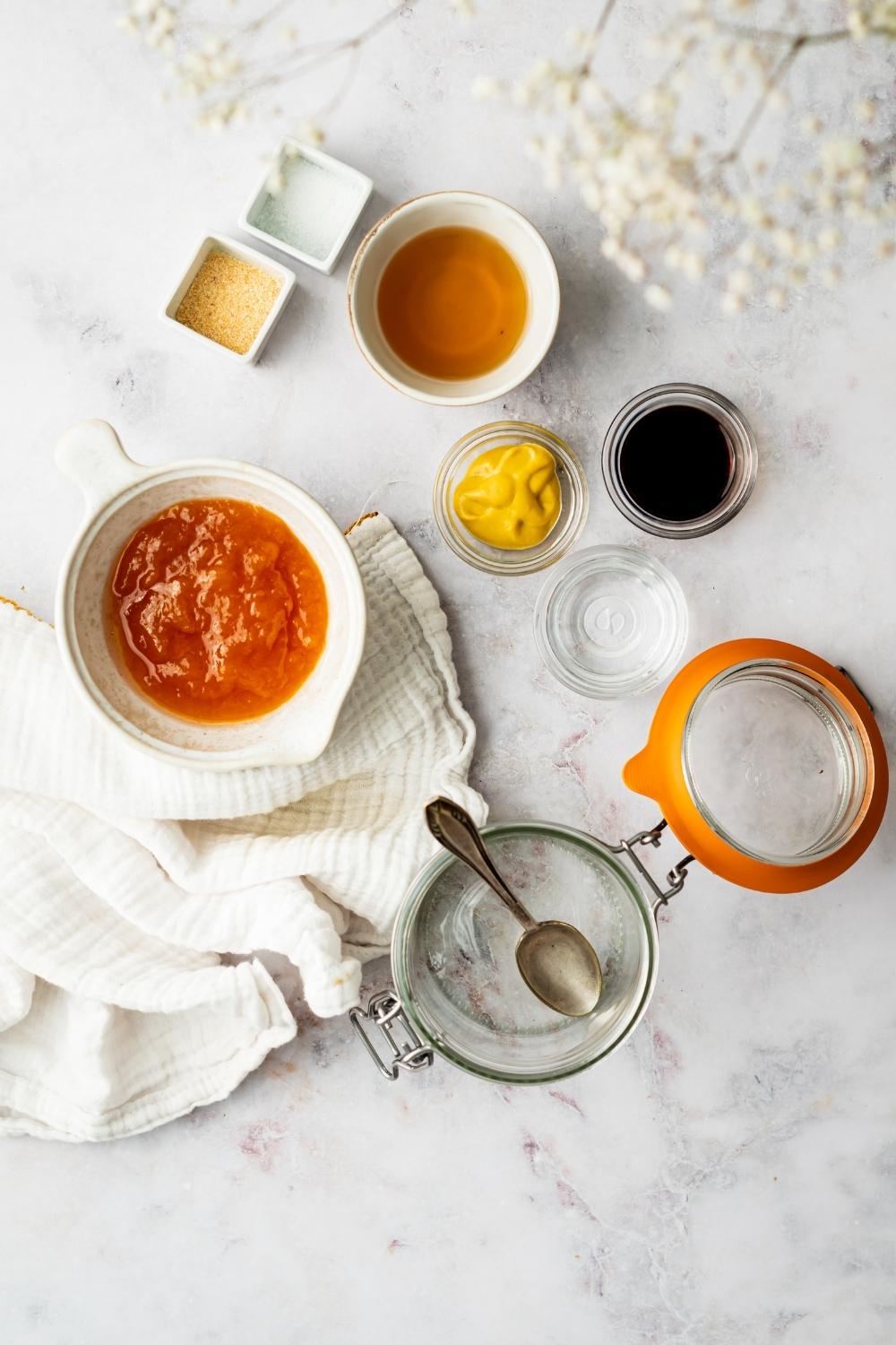 A bowl of apricot, a bowl of honey, a bowl of mustard, a bowl of soy sauce, and an empty glass jar all on a grey counter.