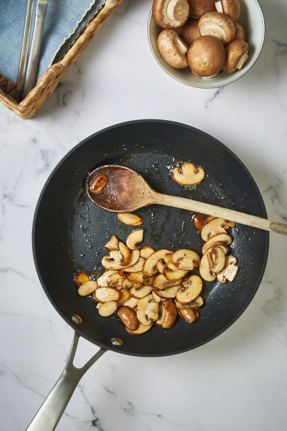 Skillet with sauteed mushrooms, a wooden spoon, and a bowl of raw mushrooms next to the skillet.