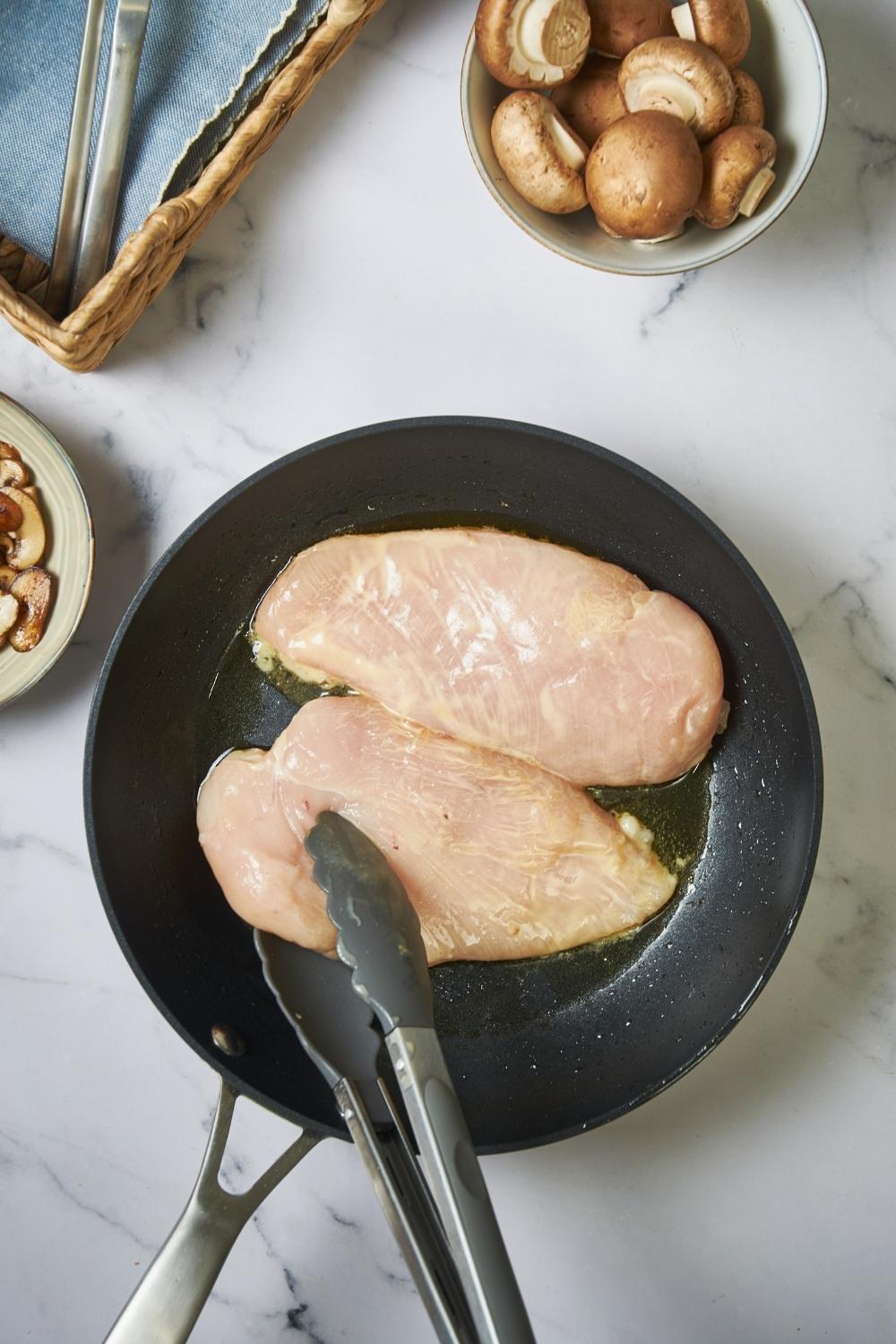 Skillet with raw chicken breast being turned over with a pair of tongs.